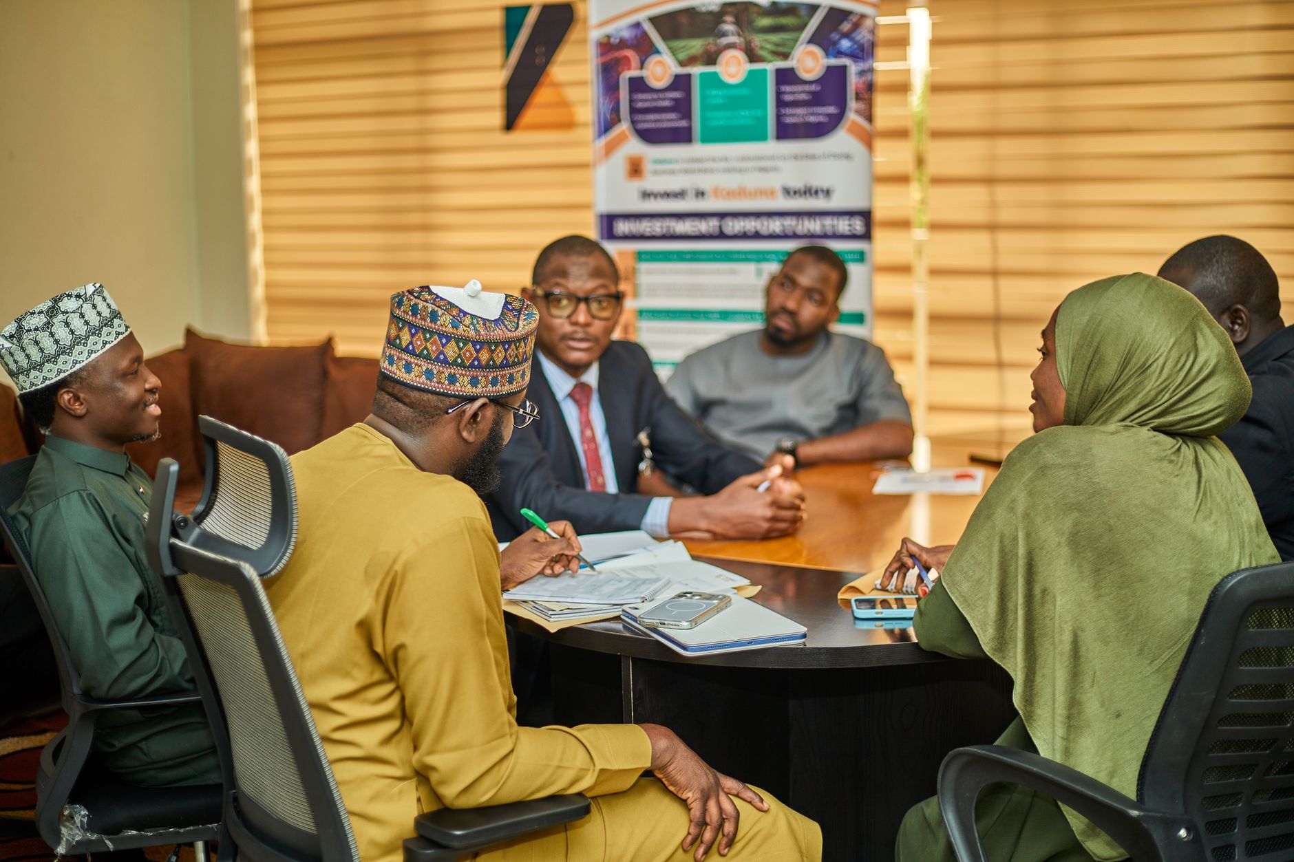 Professionals in traditional Northern Nigerian dress and a woman in hijab meeting around a boardroom table in Kaduna