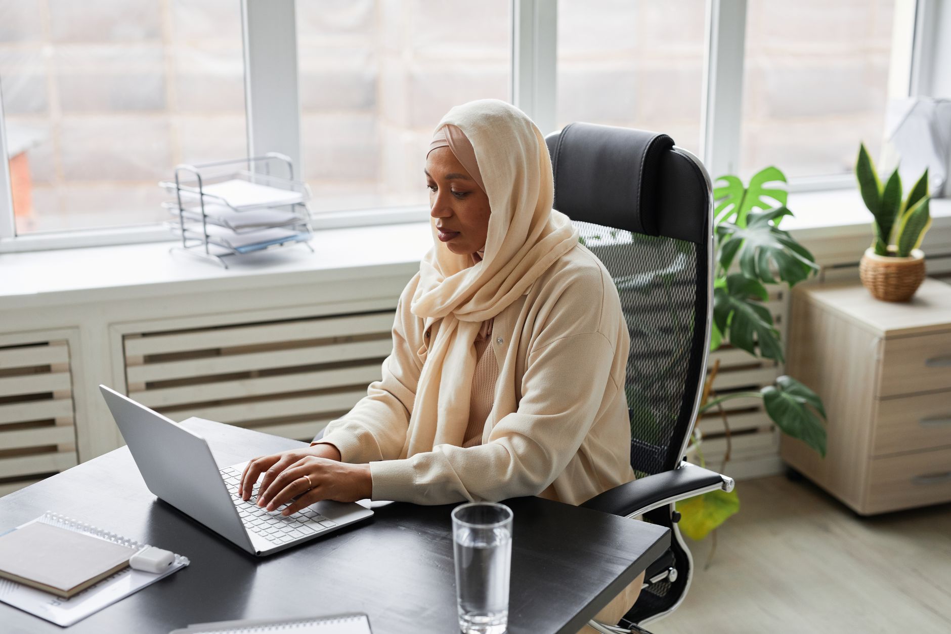 A Black woman in a hijab and glasses joining a session on her laptop at an office desk