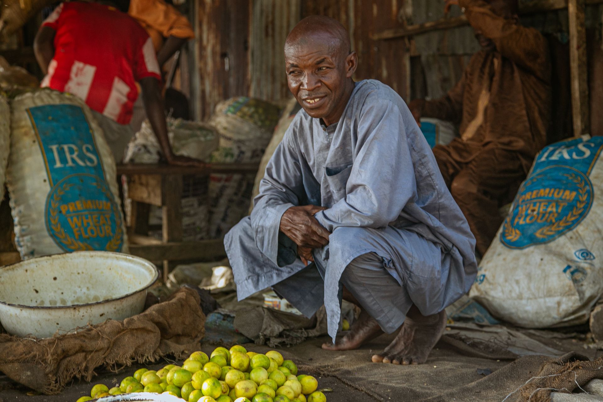 An elderly trader in his stall in a Maiduguri market, surrounded by grain sacks and produce