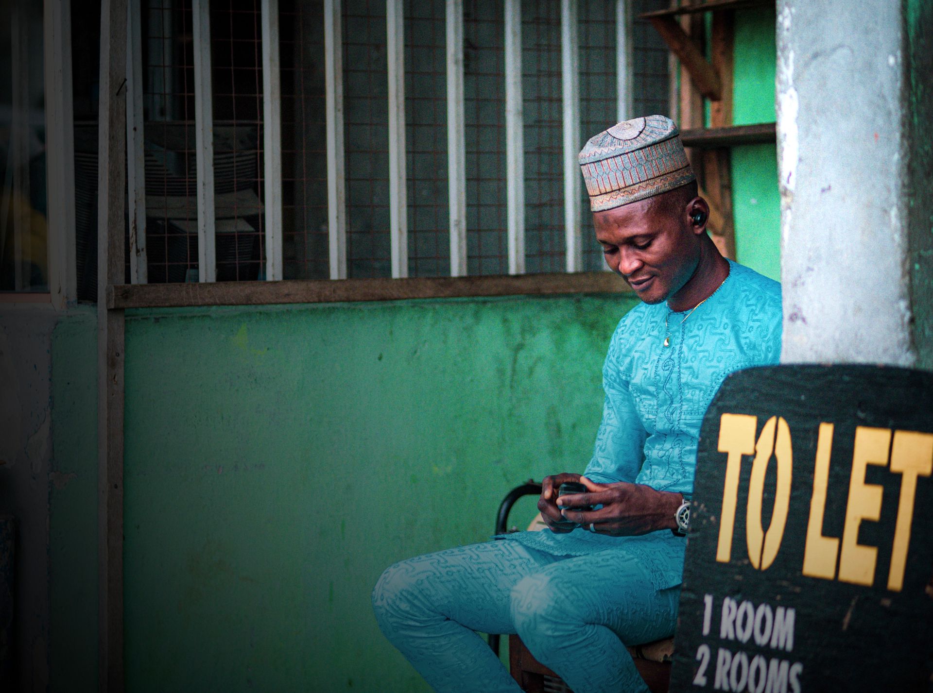 A young Nigerian man in traditional dress using his phone beside a shop front in a Nigerian town