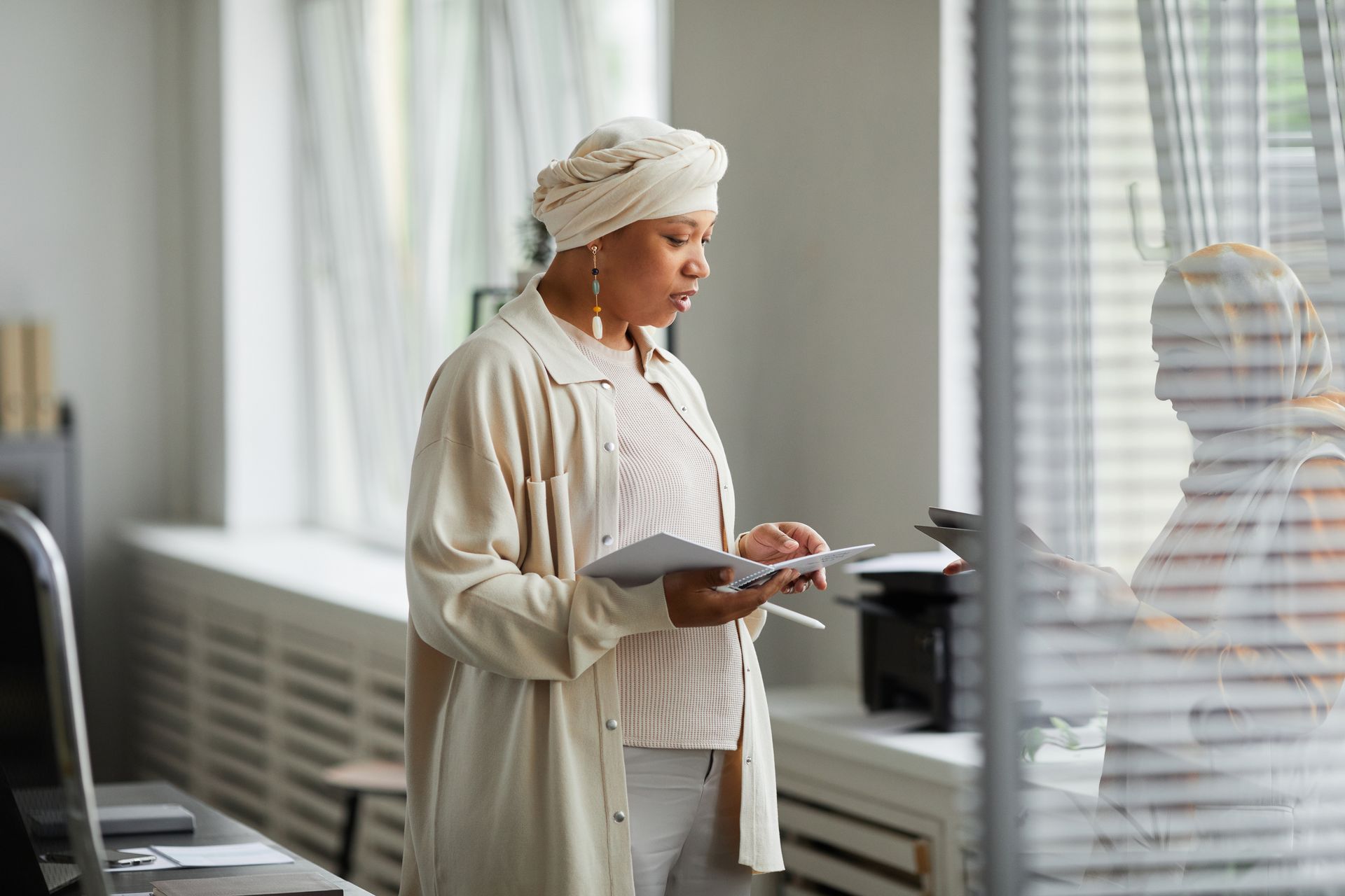 A senior Black businesswoman in a headwrap reviewing documents beside an office window